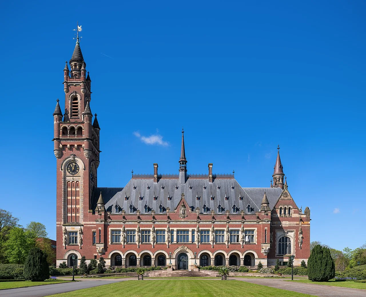 The Peace Palace in The Hague: a red-brick building with a grey slate roof and a tall clock tower at its left corner, under a clear blue sky.